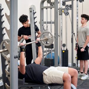 3 students are in the gym. One of them is using a barbell while the other helps direct